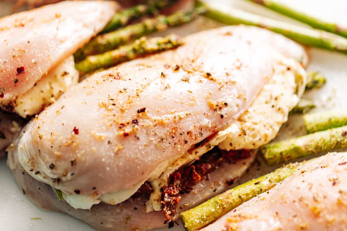 Raw stuffed chicken breasts with asparagus, mozzarella cheese and sun dried tomato strips on a white cutting board.