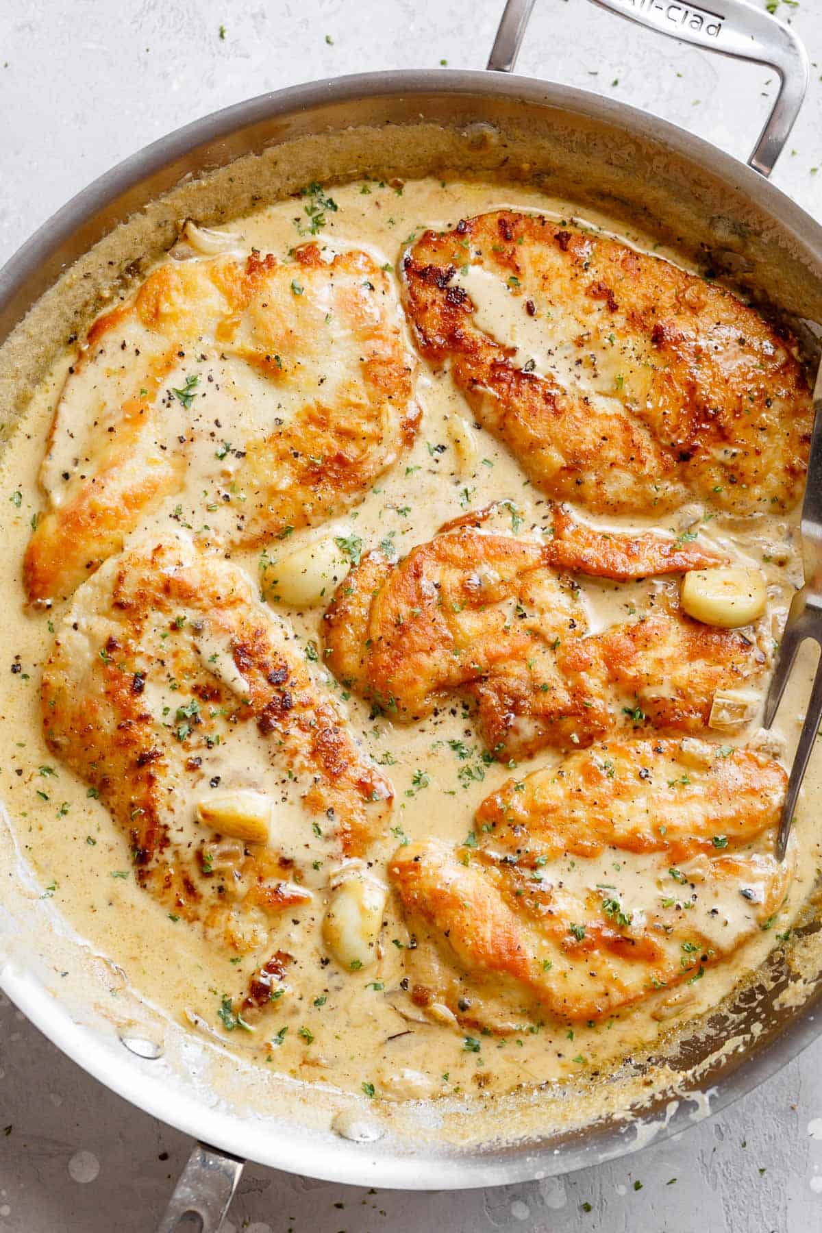 A silver frying pan of deliciously five Chicken Breasts in a garlic cream sauce with whole garlic cloves and a silver serving fork | cafedelites.com