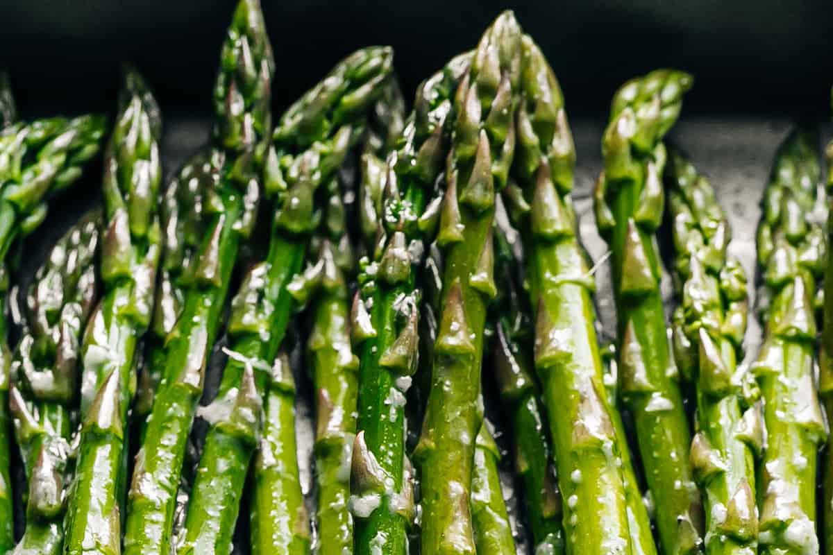 A bunch of garlic and oil coated asparagus spears on a dark grey baking sheet