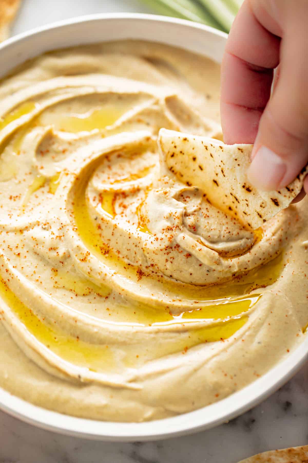 A side view image of a piece of pita bread dipped into Hummus served in a white bowl, drizzled with olive oil and topped with a sprinkle of paprika | cafedelites.com