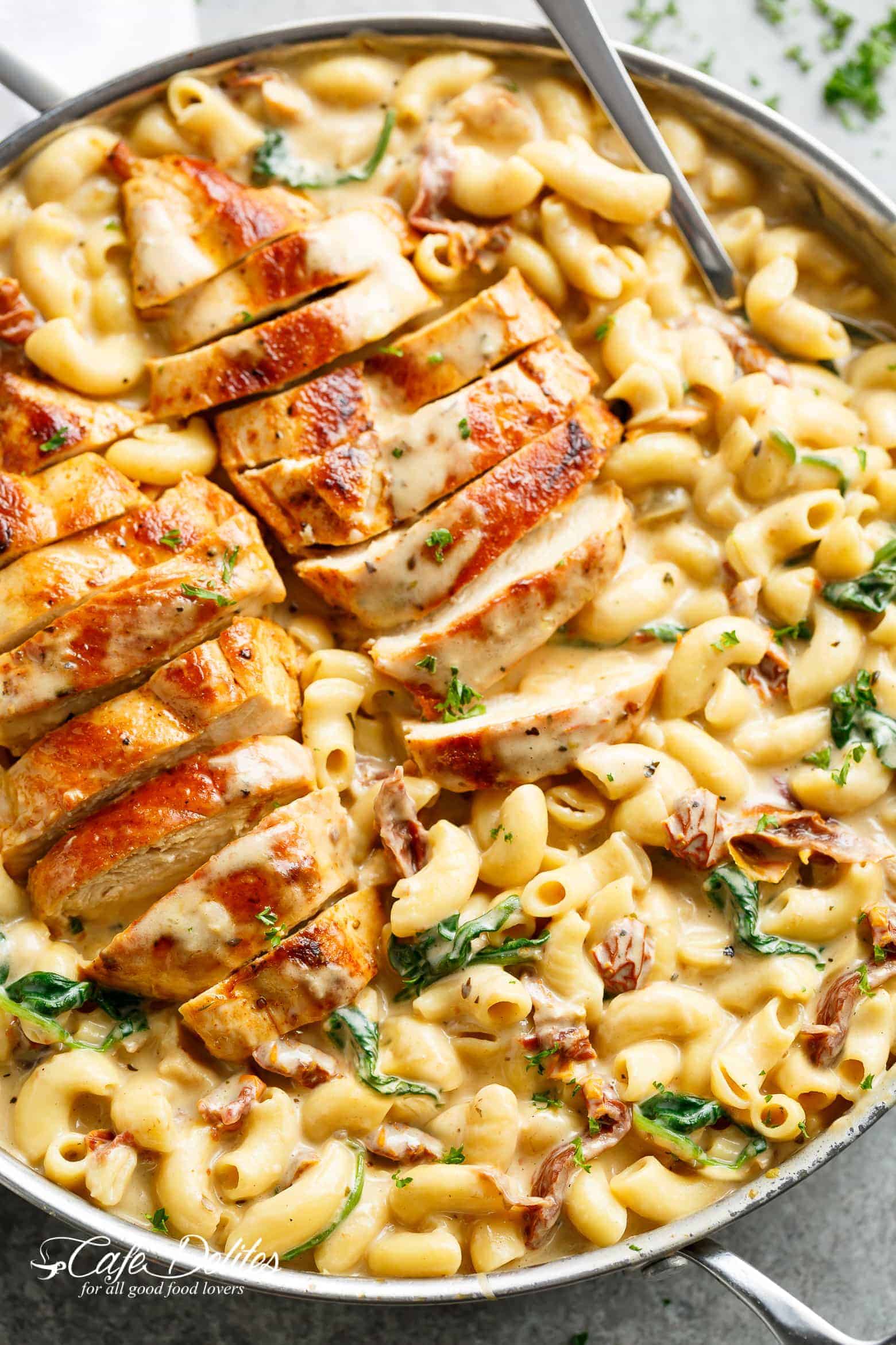 A silver frying pan filled with creamy tuscan style Mac and cheese with wilted spinach and sun dried tomato strips! Two sliced chicken breasts sit on top of Mac and cheese with a serving spoon in the pan ready to serve!