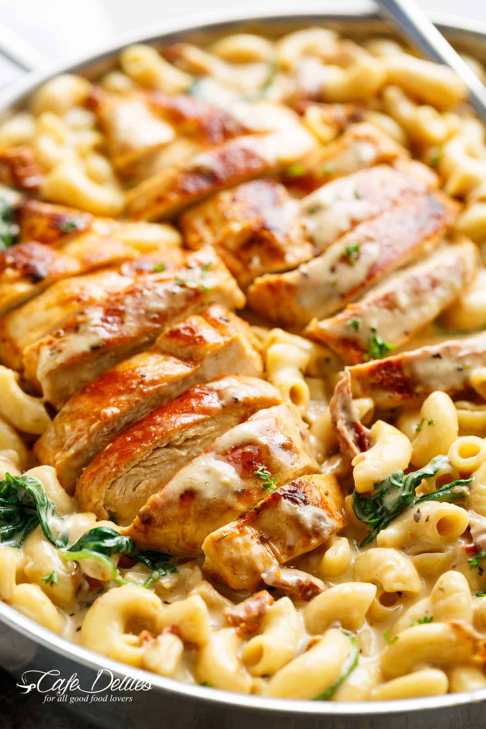 A silver frying pan filled with creamy tuscan style Mac and cheese with wilted spinach and sun dried tomato strips! Two sliced chicken breasts sit on top of Mac and cheese with a serving spoon in the pan ready to serve!
